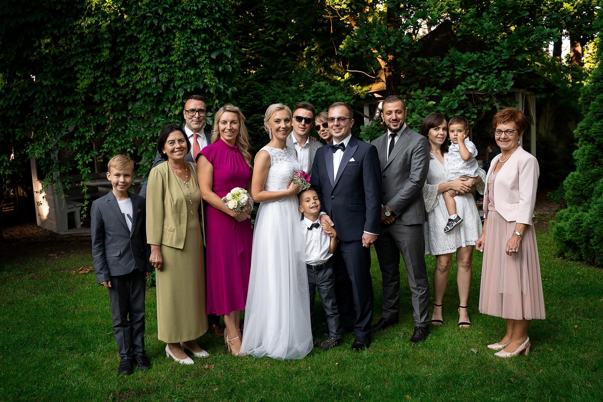 Grupowe zdjęcie ślubne w plenerze: para młoda otoczona rodziną i przyjaciółmi na tle zielonej ściany z bluszczu.