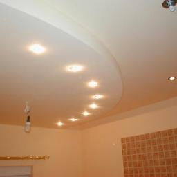 Budowa - Podwieszany sufit z oświetleniem punktowym, widoczna instalacja elektryczna z żarówką w tle, ściana wyłożona brązowymi płytkami.