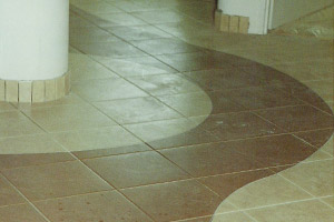 Ułożona podłoga z płytek w odcieniach beżu i brązu, z dekoracyjnym wzorem w kształcie fali, otaczająca białą kolumnę wykończoną fragmentami płytek.