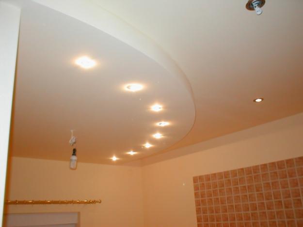 Podwieszany sufit z oświetleniem punktowym, widoczna instalacja elektryczna z żarówką w tle, ściana wyłożona brązowymi płytkami.