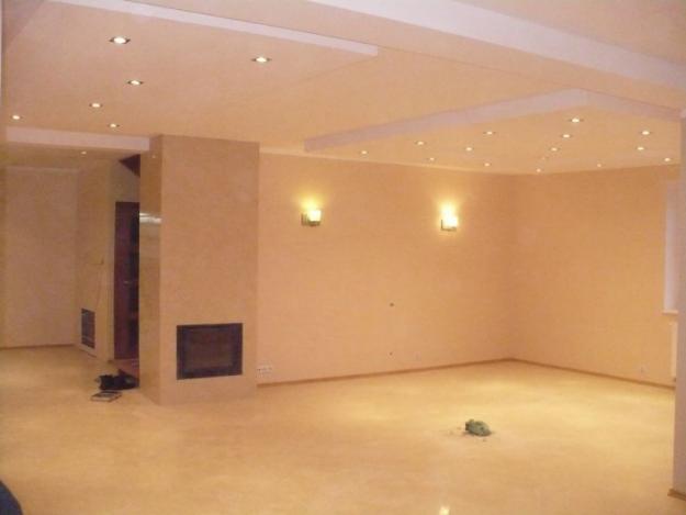 Wnętrze pokoju z beżowymi ścianami, jasną podłogą, wbudowanym kominkiem i oświetleniem punktowym w suficie.