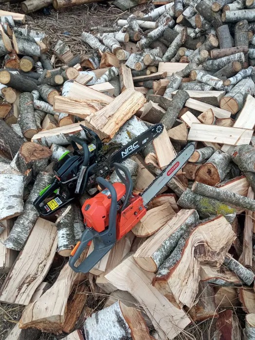 Dwie piły łańcuchowe (NAC i Oregon) leżą na stercie pociętego drewna brzozowego, przygotowanego do kominka.