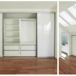 Biała zabudowa garderoby z przesuwnymi drzwiami i półkami, widok na poddasze z oknami dachowymi, drewniana podłoga.