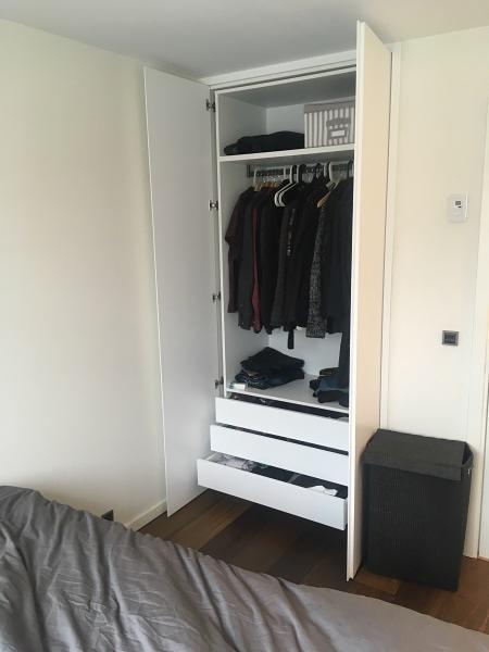 Wnętrze białej, minimalistycznej szafy wnękowej z trzema szufladami i półkami, na których wiszą ubrania w ciemnych kolorach. Częściowo otwarte drzwi szafy ukazują jej zawartość. Obok szafy stoi...