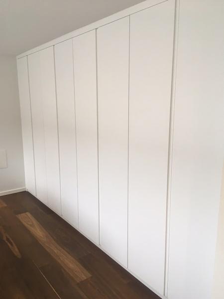 Biała zabudowa szafy wnękowej z frontami bez uchwytów, widoczna na tle parkietu z ciemnego drewna.