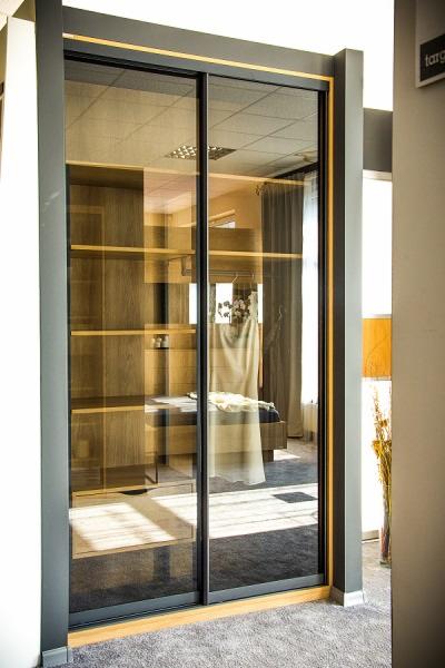 Szafa wnękowa z przesuwnymi drzwiami z ciemną ramą i lustrzanym frontem, odbijająca wnętrze sypialni z łóżkiem i garderobą z drewnianymi półkami.