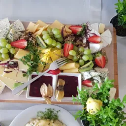 Deska serów z owocami, winogronami i konfiturami, udekorowana zielenią i figurką kurczaczka, widoczna na stole z elementami dekoracji wielkanocnych.