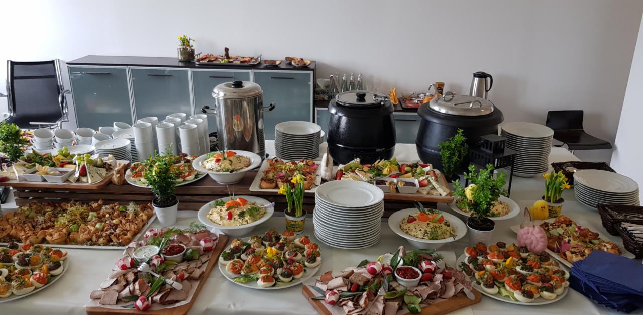 Stół cateringowy z różnorodnymi daniami: jajka faszerowane, wędliny, sałatki, sery, przekąski na pieczywie, zupy w podgrzewaczach, dekoracje z kwiatów i figurek kurczaków, biała zastawa, tło biurowe.