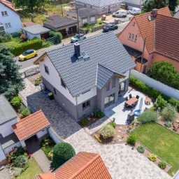 Nowoczesny dom szkieletowy z szarą dachówką, widok z lotu ptaka na posesję z patio i zadbanym ogrodem, żółty samochód w tle.