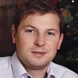 Portret mężczyzny w koszuli w delikatny wzór, z krótkimi brązowymi włosami, na tle rozmytej choinki z czerwonymi bombkami, oświetlony miękkim światłem.