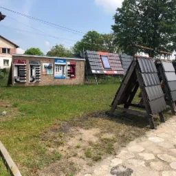 Ekspozycja różnych rodzajów pokryć dachowych: dachówki ceramiczne, blacha trapezowa oraz rynny, prezentowane na zewnątrz budynku w dzień.