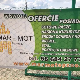 Baner reklamowy firmy MAR-MOT Sp. z o.o. z ofertą produktów, w tym gotowe pasze, nasiona kukurydzy, środki ochrony roślin, węgiel, stal i pokrycia dachowe, umieszczony na ogrodzeniu z siatki.