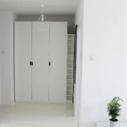 Białe, minimalistyczne wnętrze z błyszczącą, jednolitą podłogą, trzydrzwiową szafą w zabudowie, geometrycznymi lampami i rośliną doniczkową w metalowej osłonce.
