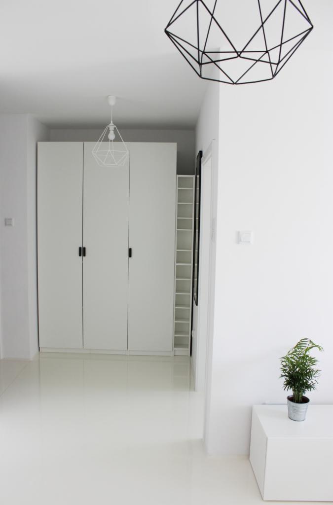 Białe, minimalistyczne wnętrze z błyszczącą, jednolitą podłogą, trzydrzwiową szafą w zabudowie, geometrycznymi lampami i rośliną doniczkową w metalowej osłonce.