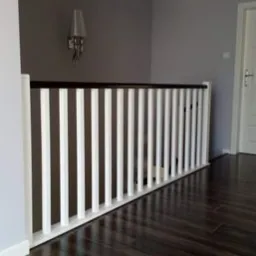 Biała balustrada z ciemnobrązowym poręczą na piętrze, widok z dołu na ciemnej podłodze, lampa ścienna w tle.