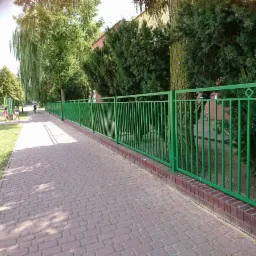 Długie, zielone ogrodzenie z pionowych prętów i dekoracyjnymi rombami, zamontowane na ceglanym murku, oddziela chodnik z kostki brukowej od terenu zielonego z placem zabaw w tle.