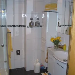 Jasna łazienka z białymi płytkami i czarną listwą dekoracyjną, kabina prysznicowa po lewej, biała szafka z umywalką i bukietem słoneczników, czarna podłoga z szarą wycieraczką.