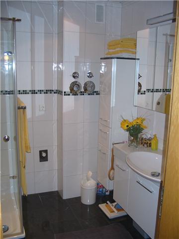 Jasna łazienka z białymi płytkami i czarną listwą dekoracyjną, kabina prysznicowa po lewej, biała szafka z umywalką i bukietem słoneczników, czarna podłoga z szarą wycieraczką.