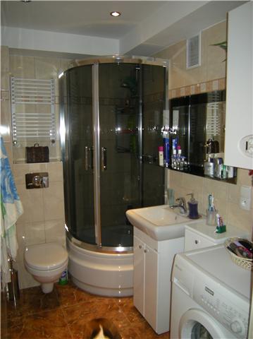 Ukończona łazienka z kabiną prysznicową, umywalką z szafką, toaletą i pralką. Na ścianach beżowe płytki, na podłodze brązowe. Widoczny grzejnik drabinkowy i lustro z półką na kosmetyki.