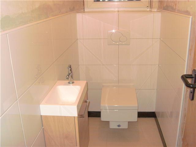 Wykończona łazienka z beżowymi płytkami ułożonymi w karo na ścianie, wisząca toaleta i umywalka z szafką z jasnego drewna.