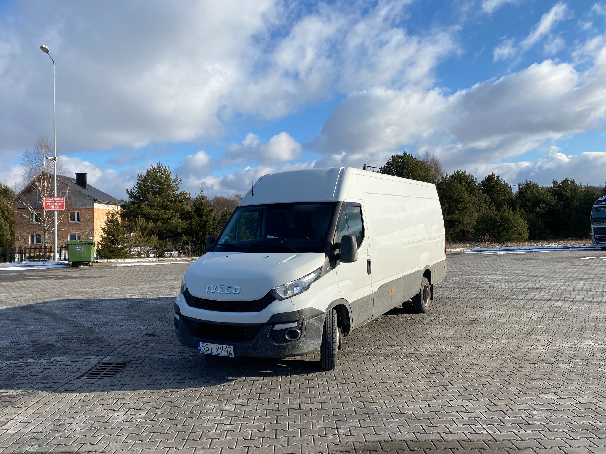 Biały bus Iveco Daily zaparkowany na parkingu z kostki brukowej w słoneczny dzień, widoczne budynki w tle i błękitne niebo z chmurami.
