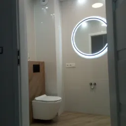 Nowoczesna łazienka z podwieszaną toaletą, okrągłym lustrem z podświetleniem LED i płytkami imitującymi drewno na podłodze, widziana zza uchylonych drzwi.