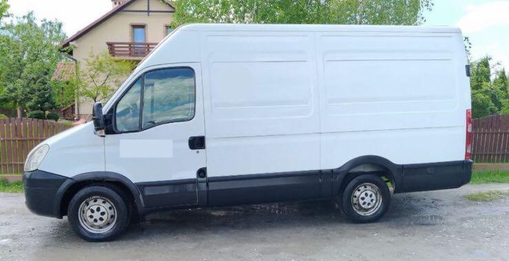 Biały samochód dostawczy z wysokim dachem na tle domu jednorodzinnego z brązowym dachem i drewnianego płotu.