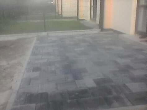 Ułożona szara kostka brukowa na posesji, widoczny fragment budynku i trawnika w tle, zacieranie fug w trakcie prac.