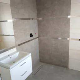 Nowoczesna łazienka w trakcie wykańczania, beżowe płytki z metalowymi listwami, biała szafka z umywalką, odpływ liniowy w podłodze.