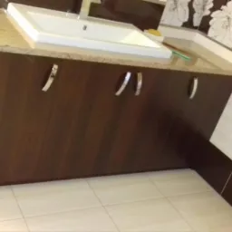 Nowoczesna szafka pod umywalkę z ciemnego drewna z jasnym blatem, srebrne uchwyty, biała umywalka, jasne płytki podłogowe, dekoracyjne płytki ścienne z motywem kwiatowym w tle.