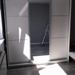 Biała szafa przesuwna z lustrem w centralnej części, widoczna drabina malarska w głębi pokoju, podłoga z paneli imitujących drewno.