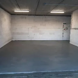 Świeżo wylana, gładka, szara posadzka w surowym pomieszczeniu z białymi ścianami z betonu komórkowego i betonowym stropem, oświetlonym dwiema lampami jarzeniowymi.