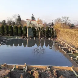 Świeżo wylana, gładka, szara powierzchnia fundamentu w drewnianym szalunku, z odbiciem drzewa i nieba, otoczona ziemią i elementami krajobrazu.