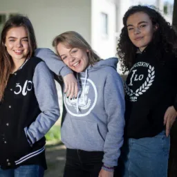 Trzy uśmiechnięte nastolatki w bluzach i kurtce z logo szkoły, stojące na tle budynku.