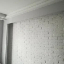 Biała ściana z cegły pomalowana na biało, widoczna listwa sufitowa i fragment ramy okiennej, minimalistyczny wystrój wnętrza.