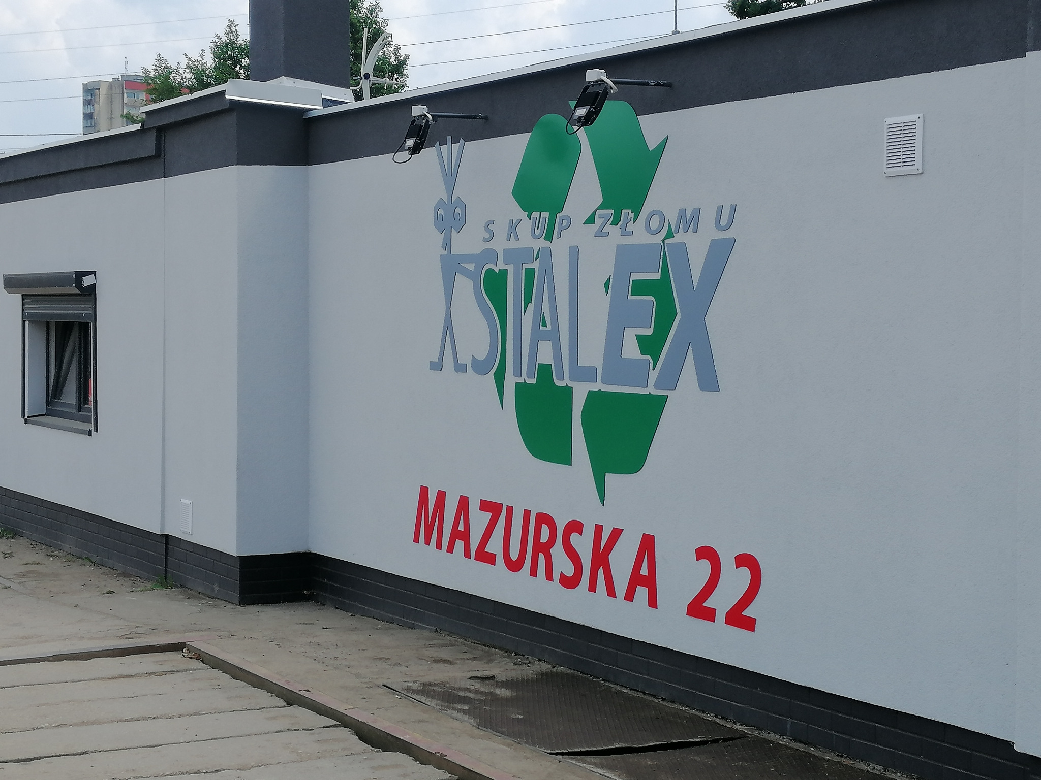Elewacja budynku z dużym logo 'Skup Złomu Xstalex' wkomponowanym w symbol recyklingu i adresem 'Mazurska 22', oświetlona dwoma reflektorami.