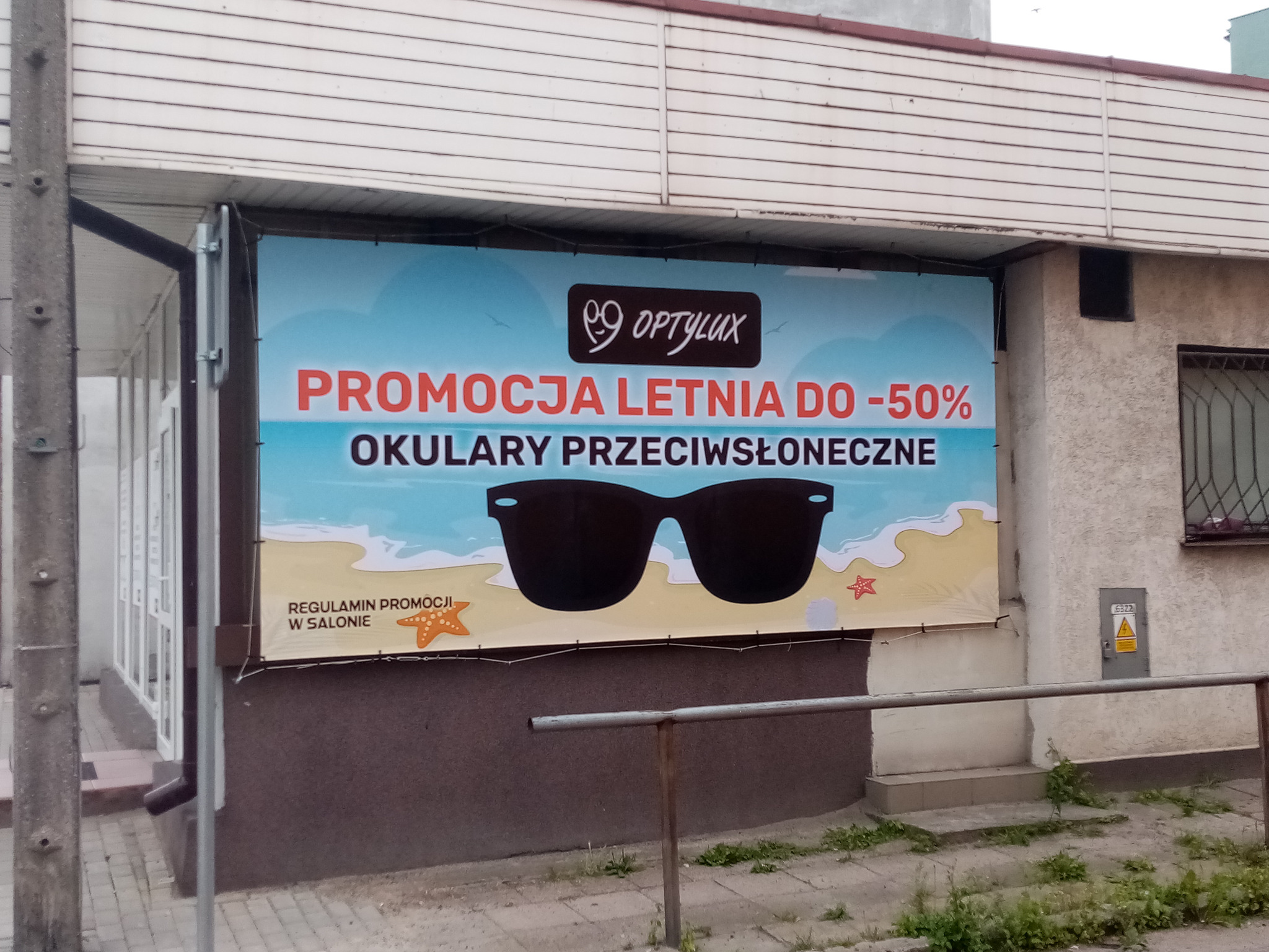 Baner reklamowy salonu optycznego Optylux z letnią promocją okularów przeciwsłonecznych, zawieszony na budynku, z grafiką plaży i okularów.