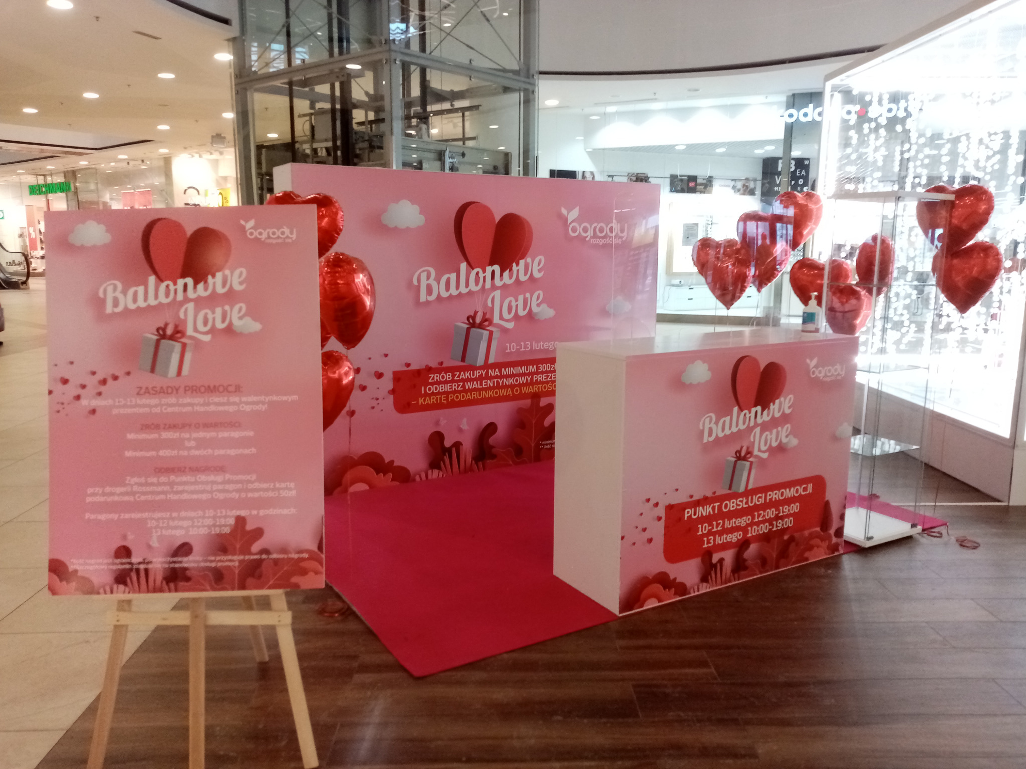 Walentynkowa strefa promocyjna 'Balonove Love' w centrum handlowym Ogrody, z czerwonymi balonami w kształcie serc, plakatami informacyjnymi na sztalugach i stanowiskiem obsługi promocji.