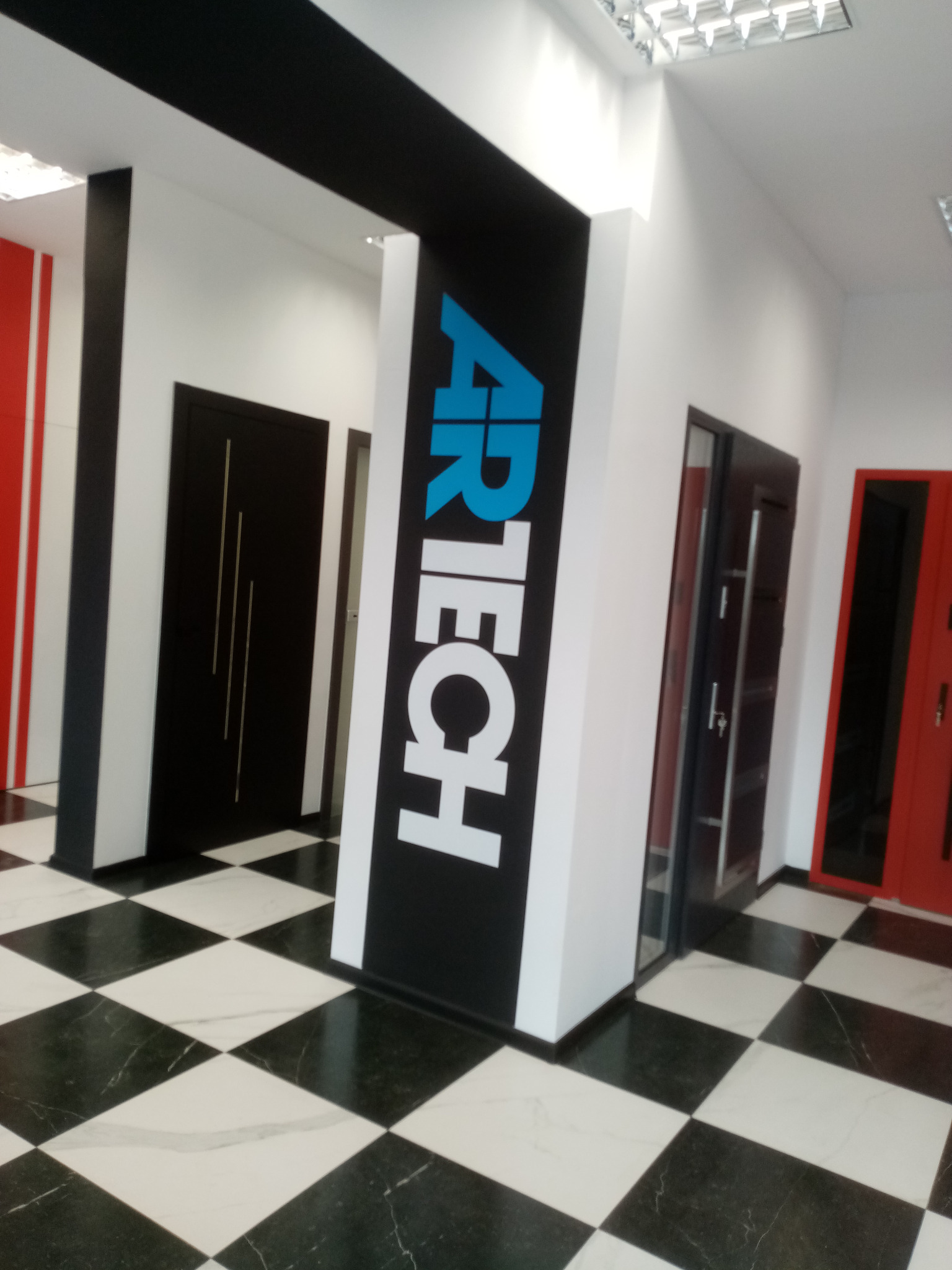 Wnętrze sklepu z drzwiami: widoczne drzwi w kolorach czerni i czerwieni, logo 'AIRTECH' na czarnym panelu, podłoga w czarno-białą szachownicę.