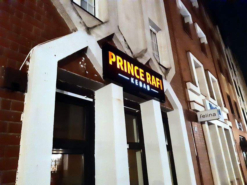 Oświetlony szyld 'Prince Rafi Kebab' nad wejściem do lokalu w budynku z cegły i białymi detalami architektonicznymi, widoczny również mniejszy szyld 'felina' obok.