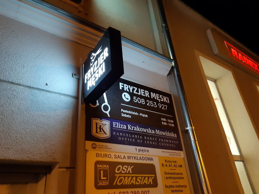 Zewnętrzny szyld salonu fryzjerskiego męskiego z podświetlanym logo i informacjami kontaktowymi, zawieszony na elewacji budynku obok szyldów innych firm, widoczny w nocnym oświetleniu.