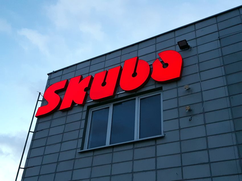 Czerwony, podświetlany szyld z nazwą firmy 'SKUBA' umieszczony na szarym budynku z widocznym oknem i niebieskim niebem w tle.