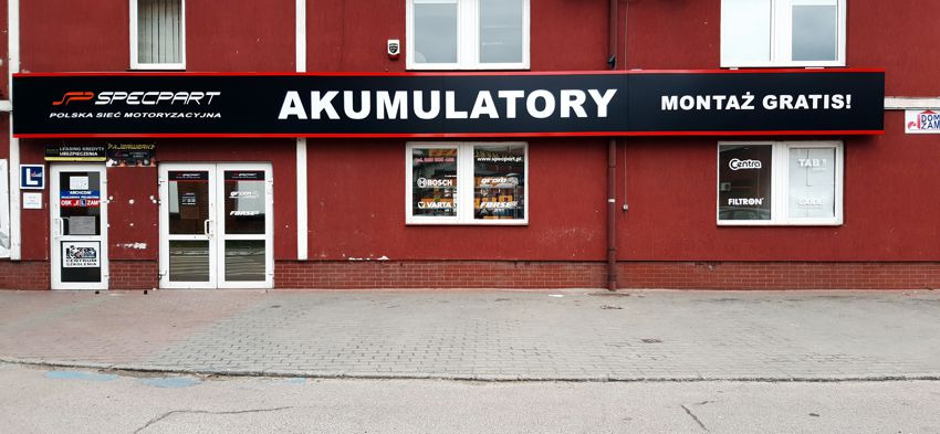 Fasada budynku sklepu motoryzacyjnego Specpart z szyldem 'Akumulatory Montaż Gratis!' i oknami z reklamami marek akumulatorów.