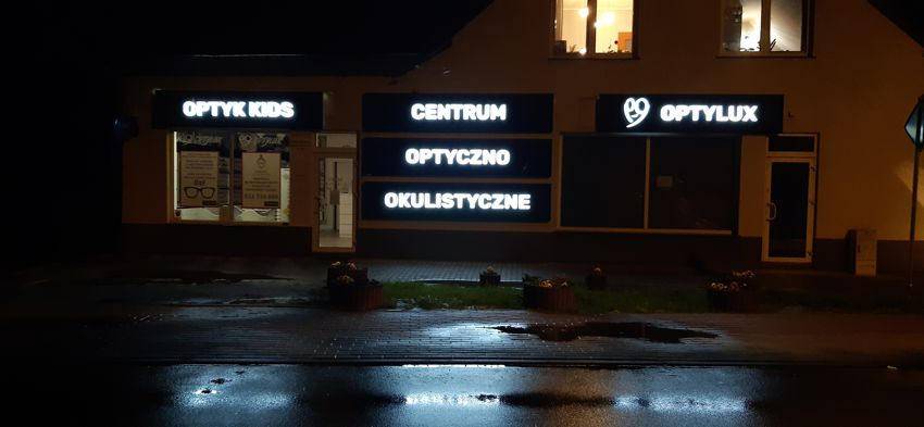 Fasada budynku z trzema szyldami: 'OPTYK KIDS', 'CENTRUM OPTYCZNO OKULISTYCZNE' i 'OPTYLUX', oświetlona nocą, z mokrym chodnikiem w kadrze.