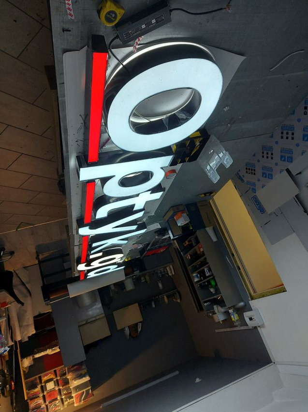 Podświetlany szyld z białymi i czerwonymi literami, widoczny w warsztacie z narzędziami i materiałami, zasilacz na górze.