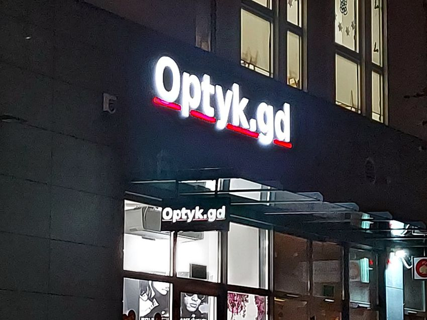 Oświetlony szyld optyka 'Optyk.gd' z czerwonymi akcentami, widoczny nad wejściem do sklepu nocą.