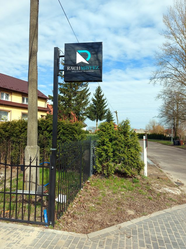Czarny, kwadratowy szyld z logo i nazwą 'RACHMISTRZ Twoje biuro rachunkowe' umieszczony na metalowym słupku przed budynkiem i ogrodzeniem.