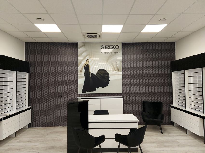 Wnętrze salonu optycznego z plakatem reklamowym Seiko, czarne fotele i szafki z okularami po bokach, ściana w ciemny wzór.