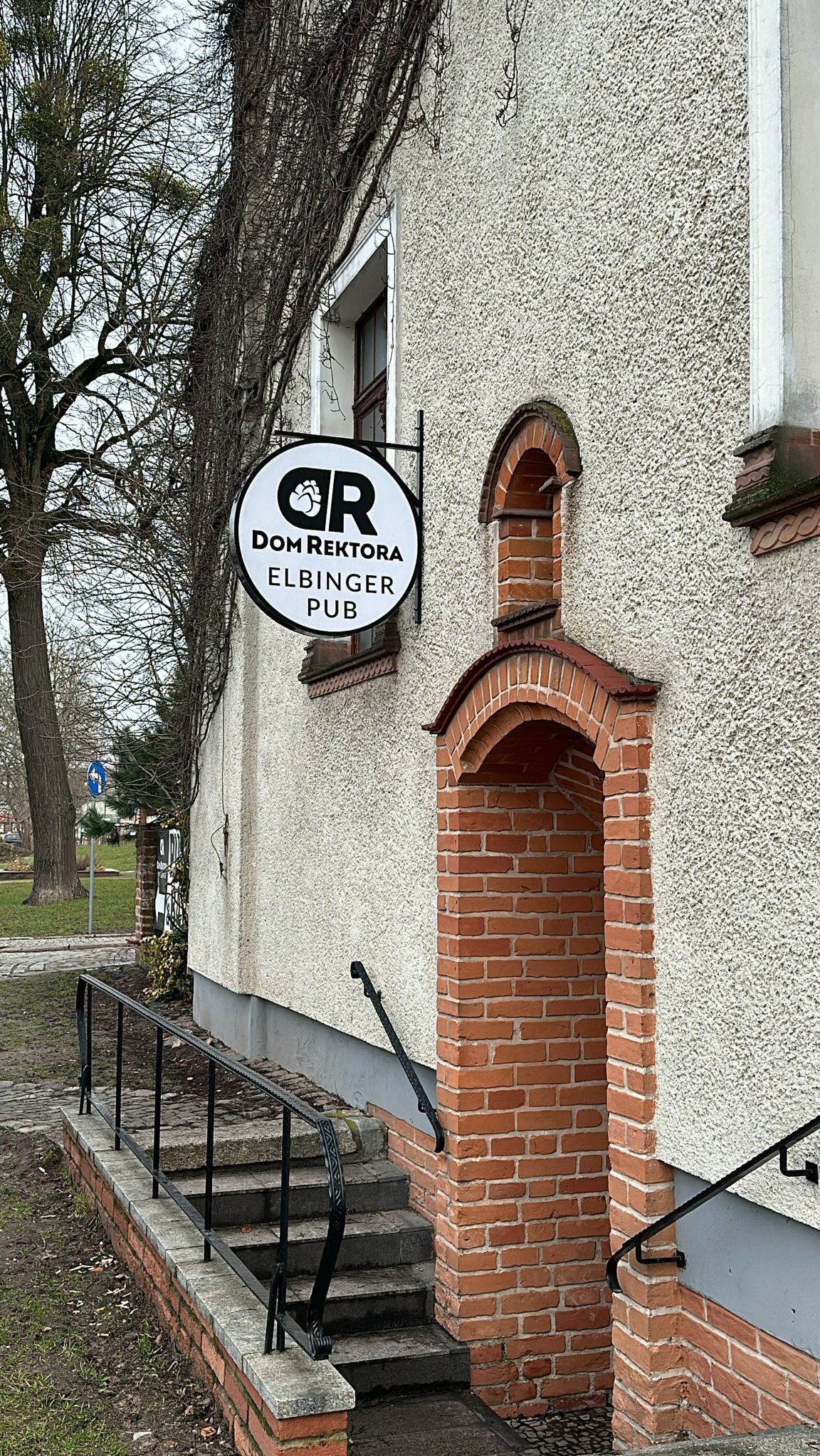 Zewnętrzny widok budynku z tablicą reklamową 'Dom Rektora Elbinger Pub' z logo niedźwiedziej łapy, ceglane wejście z łukiem, czarne metalowe balustrady schodów.