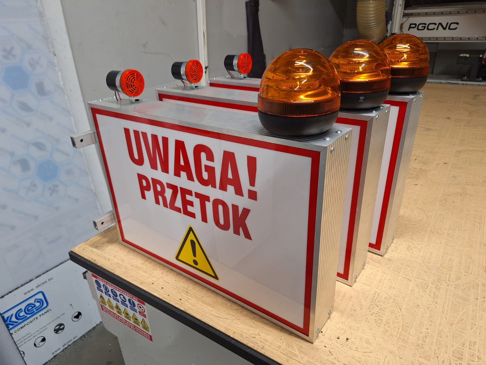Trzy identyczne, gotowe tablice ostrzegawcze z napisem 'UWAGA! PRZETOK', żółtym trójkątem ostrzegawczym, pomarańczowymi lampami ostrzegawczymi i syrenami alarmowymi, stojące na drewnianym blacie.